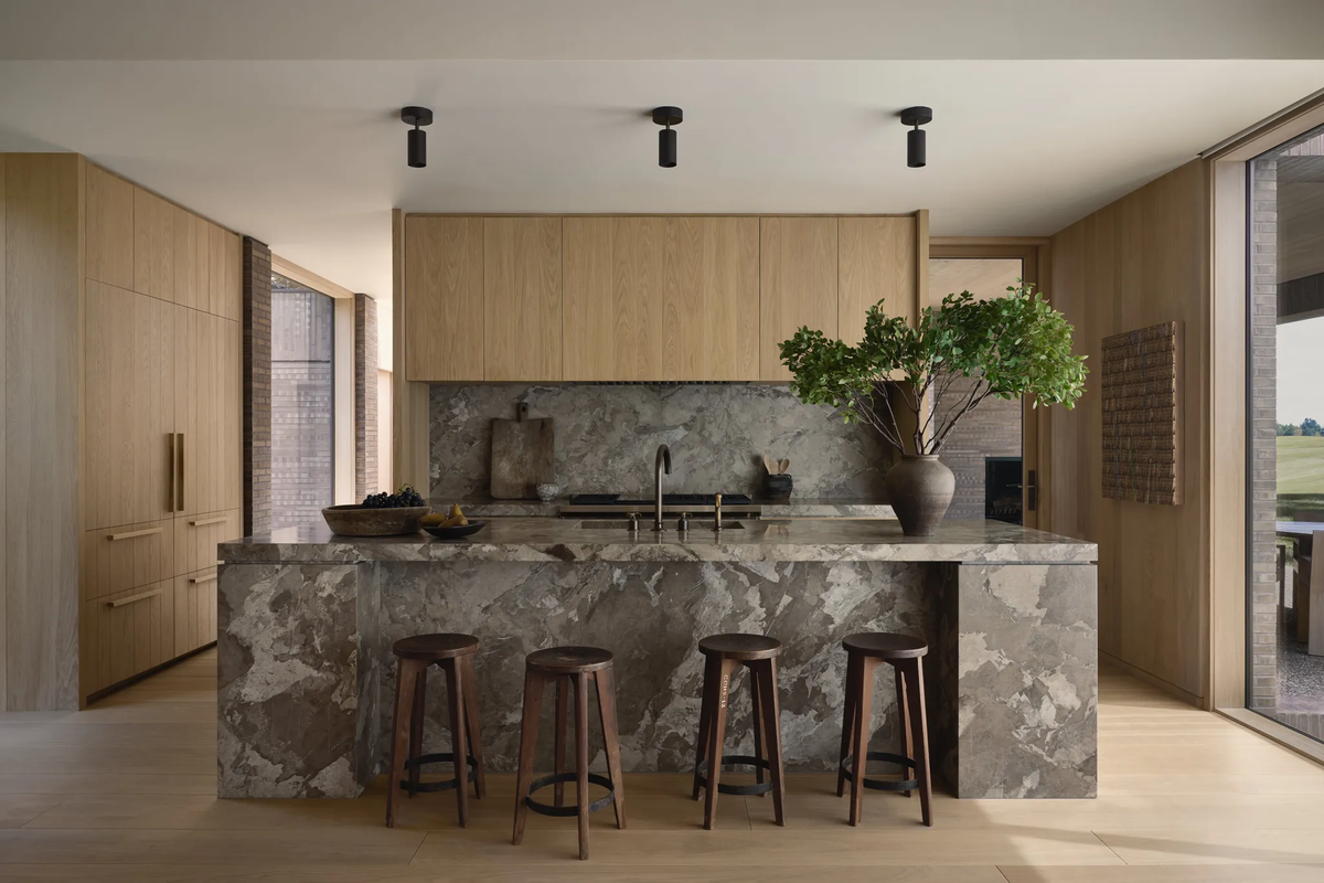 A modern kitchen with light wood cabinets, a marble island with three dark stools, a large vase of green branches, built-in appliances, and Long Made Co. Cove Adjustable Chunky Spot downlights; large windows bring in natural light.