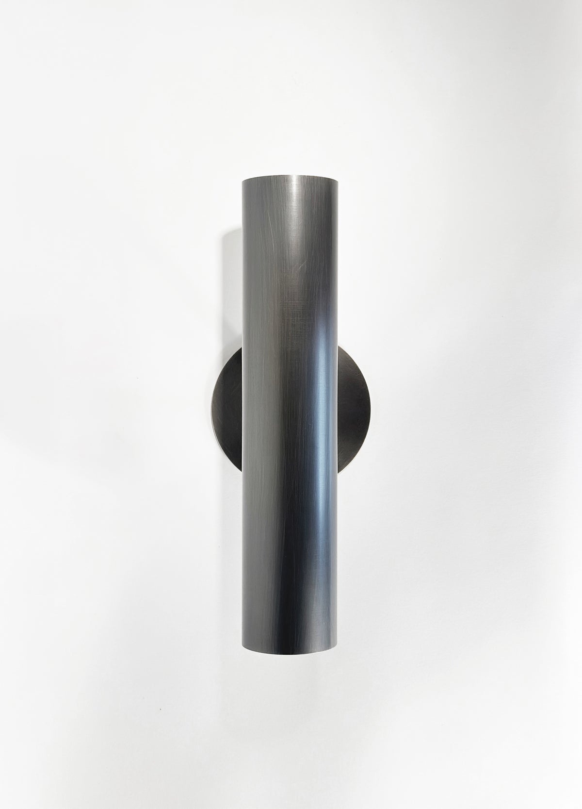 The Brass Cylinder Up-Down Sconce by Long Made Co. is a modern, cylindrical wall light with a sleek, minimalist design and round base plate, mounted vertically on a white wall.