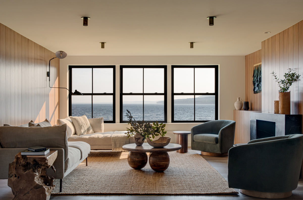 A modern living room with ocean views, neutral sofas, green armchairs, round wooden tables, a jute rug, and plant-filled minimalist decor is illuminated by the Brass Cylinder Flush Spot from Long Made Co.