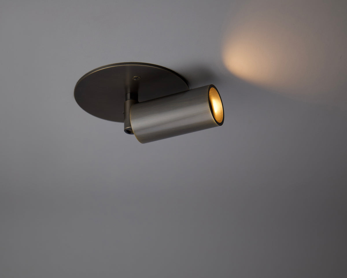 The Long Made Co. Micro Adjustable Spot, a modern cylindrical brass spotlight with a metallic finish, casts a focused warm glow onto a gray ceiling—an ideal minimalist lighting solution for any space.