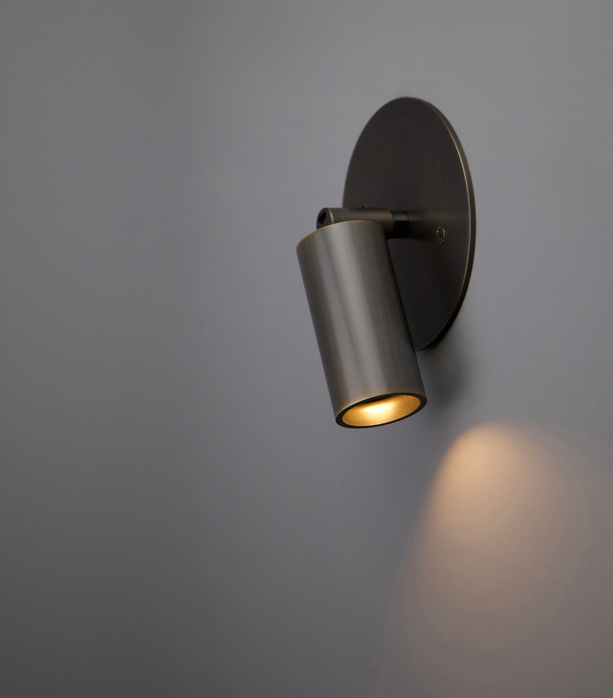 The Micro Wall Spot by Long Made Co. is a handmade brass wall sconce with an ultra-thick cylinder head, providing a warm light that softly illuminates plain grey walls.