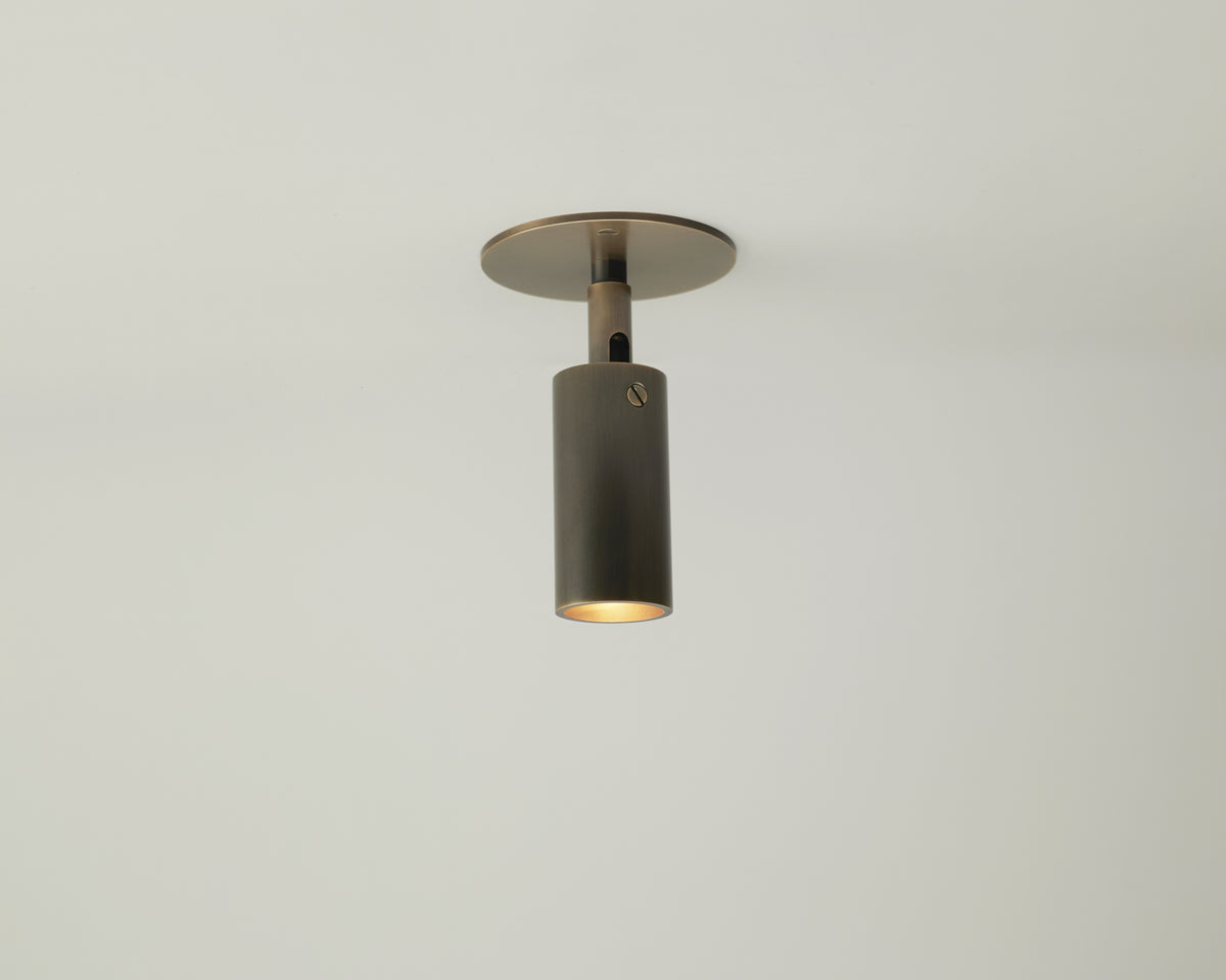 The Long Made Co. Micro Adjustable Spot, a modern cylindrical ceiling light with an ultra-thick inset cylinder head and metallic finish, is mounted on a plain white ceiling to cast soft, minimalist accent lighting downward.
