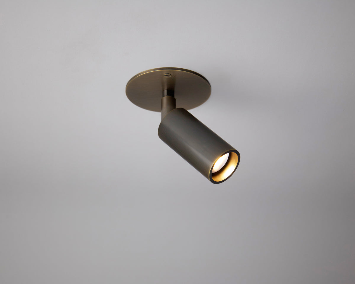 The Long Made Co. Micro Adjustable Spot is a modern cylindrical ceiling spotlight with a metallic finish on a round base, casting focused minimalist lighting downward onto a plain white ceiling.