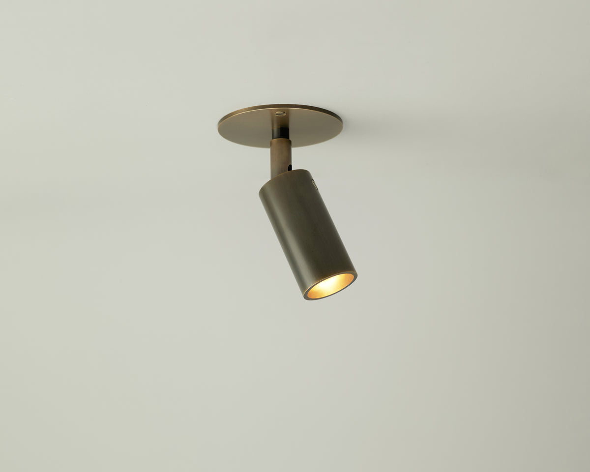 A Long Made Co. Micro Adjustable Spot, featuring a bronze finish and an ultra-thick inset cylinder head, is mounted on a plain white ceiling, angled downward and switched on.