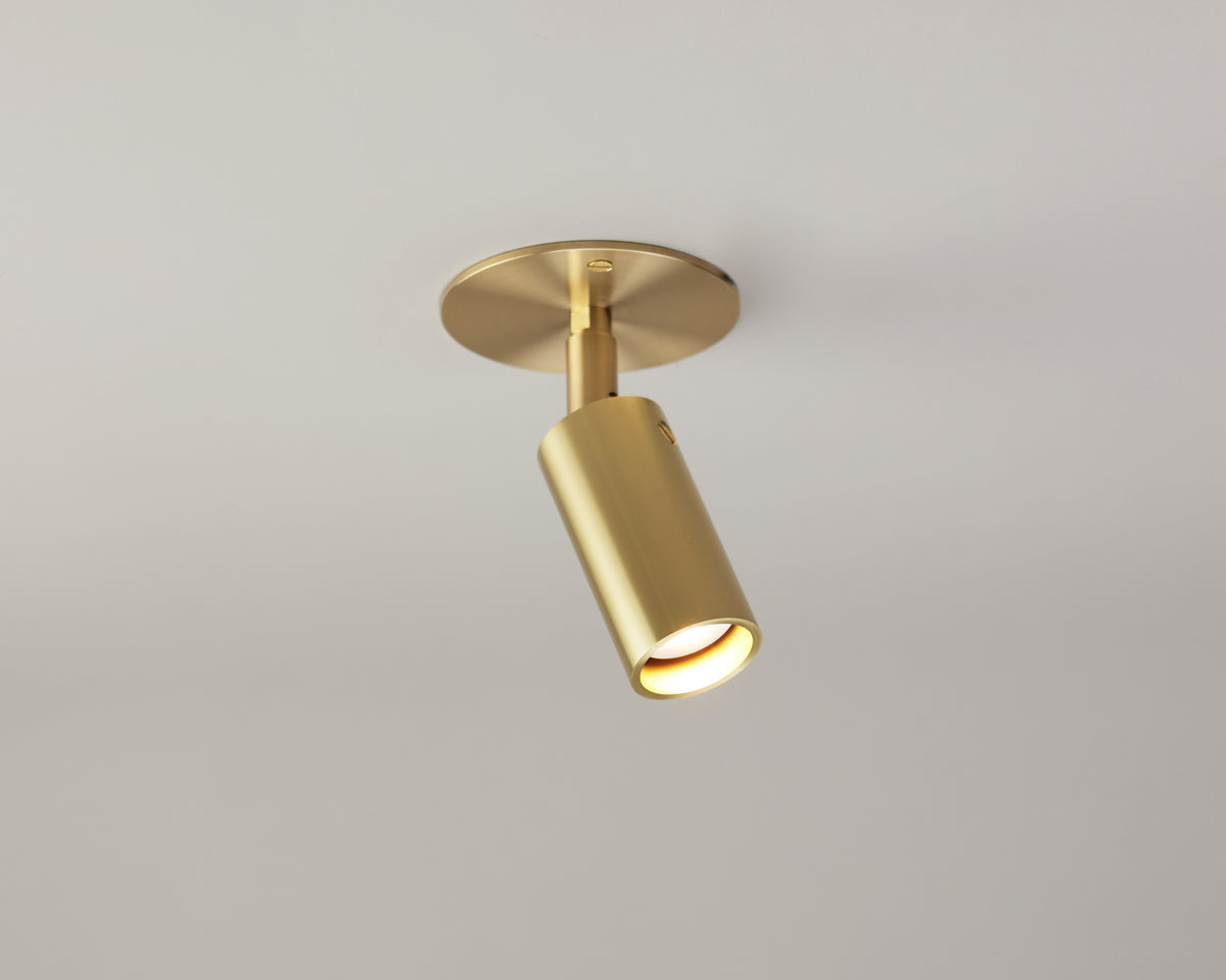 A gold Micro Adjustable Spot cylinder from Long Made Co. is mounted on a round base against a plain white ceiling, casting focused, minimalist accent lighting downward.