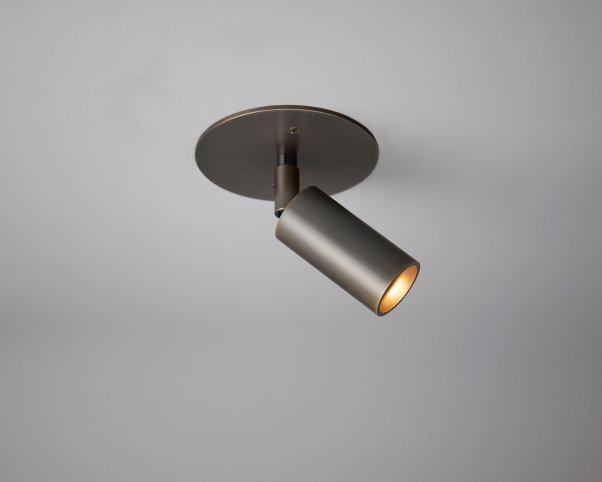 The Long Made Co. Micro Adjustable Spot is a modern, wall-mounted cylindrical spotlight with a metallic finish and minimalist design, attached to a circular base, emitting a warm focused beam from the micro spot series.