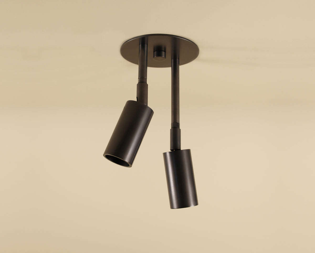 The Long Made Co. Micro Double Drop Spot is a minimalist brass fixture with two adjustable black cylindrical spotlights on a round base, shown against a plain beige background.