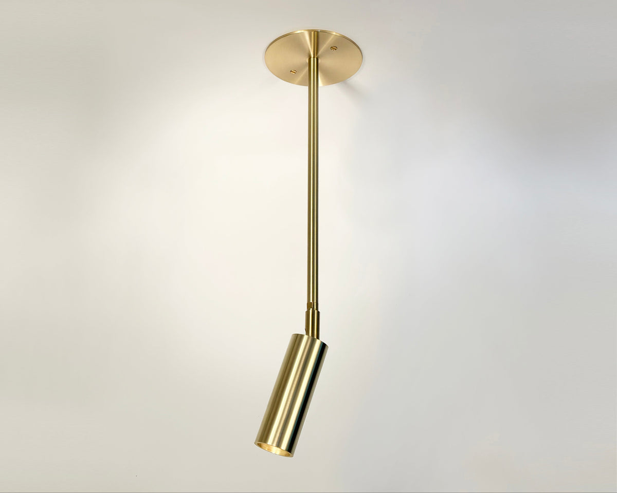 The Micro Drop Spot - w/ Long Shade by Long Made Co. is a modern brass fixture with a cylindrical spotlight on a round base, providing minimalist lighting when mounted on a white ceiling.