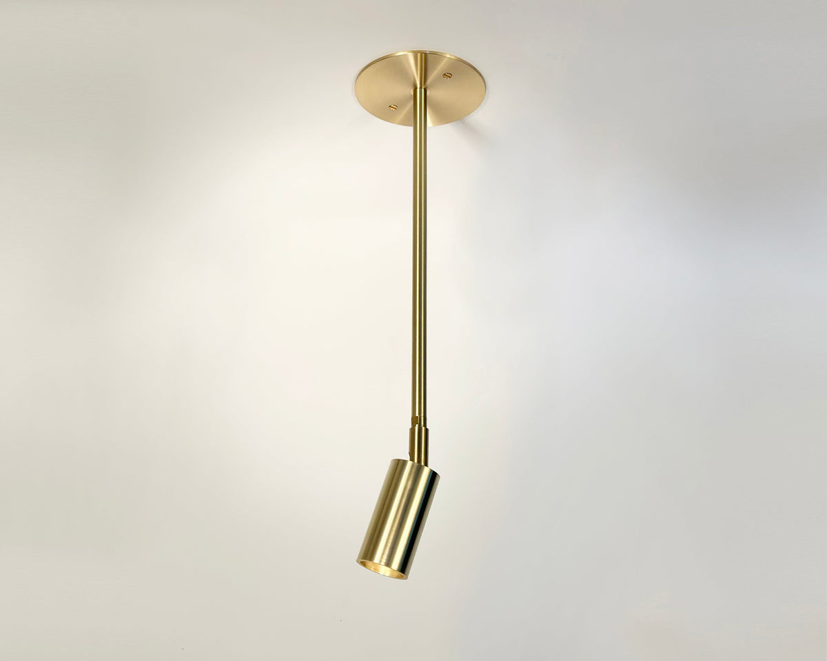 The Long Made Co. Micro Drop Spot is a minimalist brass ceiling light with an adjustable cylindrical head and sleek rod, casting soft shadows on plain walls—a premium choice for modern interiors.