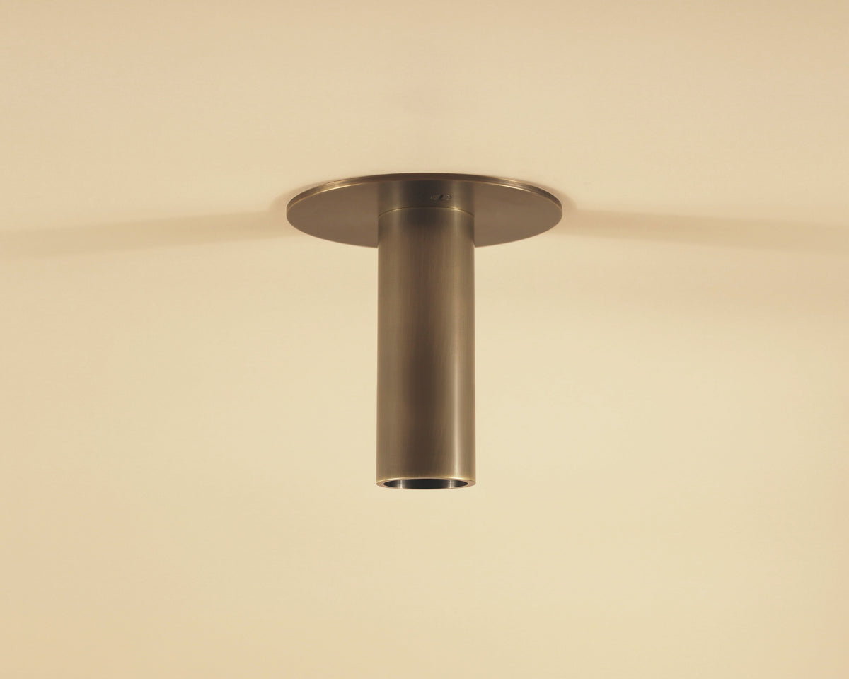 The Micro Flush Spot with Long Shade by Long Made Co. is a cylindrical, bronze-colored ceiling light extending from a flat base on a beige ceiling, softly illuminating the space.