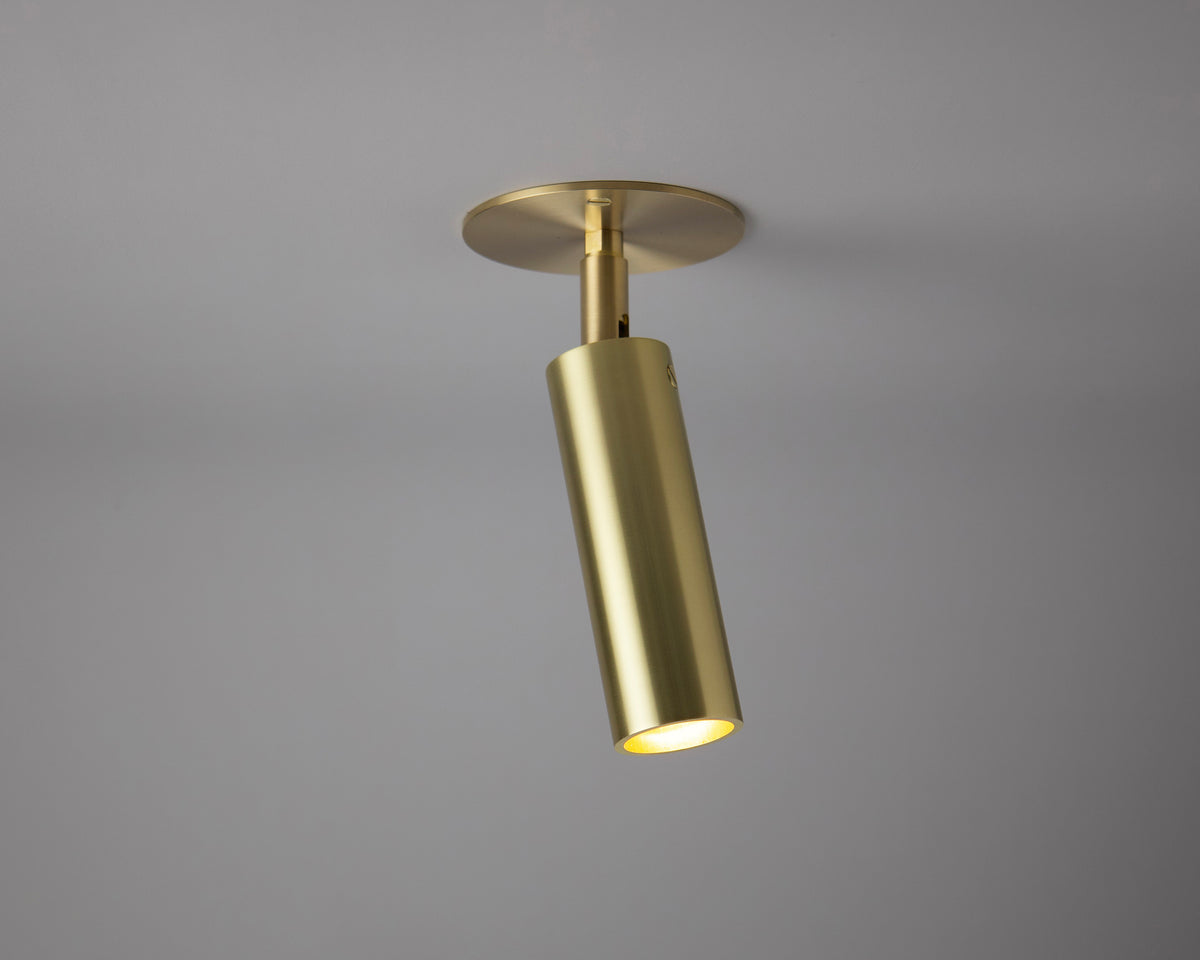 The Micro Adjustable Spot - w/ Long Shade by Long Made Co., a modern gold cylindrical ceiling spotlight with a circular base, is mounted on a light gray ceiling and angled downward to illuminate a small area below.