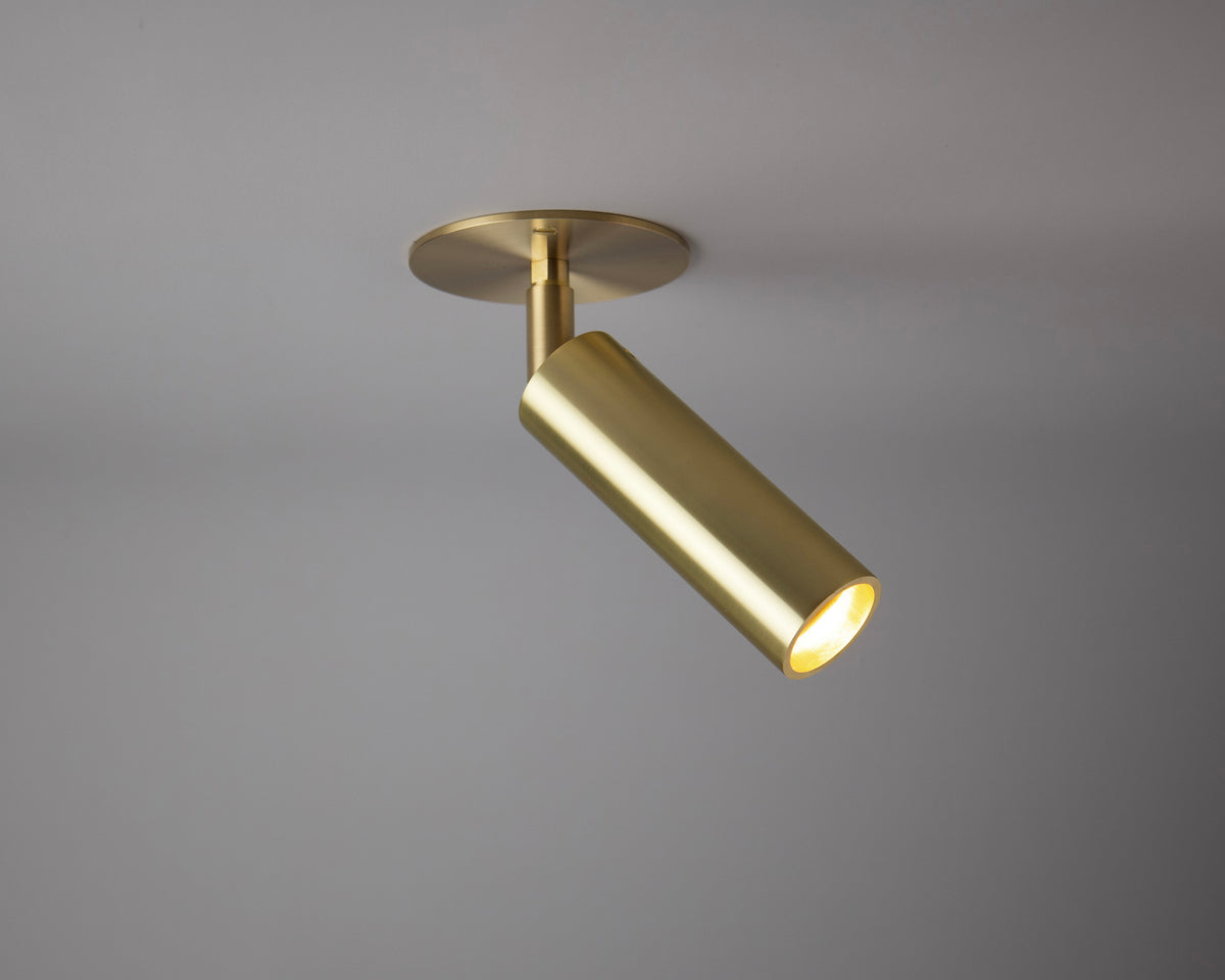 The Micro Adjustable Spot with Long Shade by Long Made Co. is a modern gold ceiling spotlight on a circular base, angled downwards for minimalist lighting that highlights part of a plain gray ceiling.