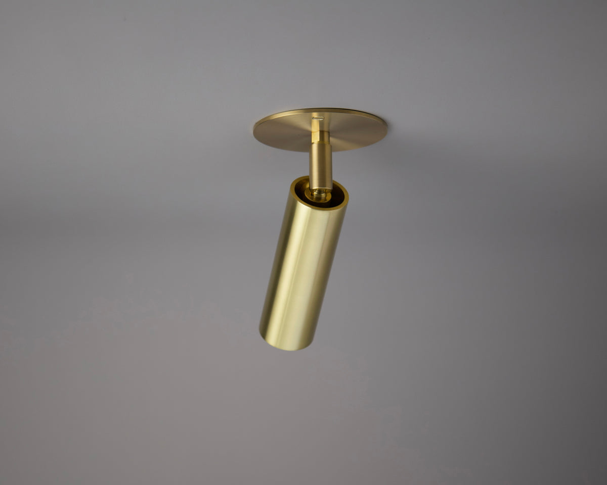 The Long Made Co. Micro Adjustable Spot - w/ Long Shade is a gold cylindrical ceiling light with an adjustable head on a round base, shown against a grey background—ideal for fans of minimalist lighting.