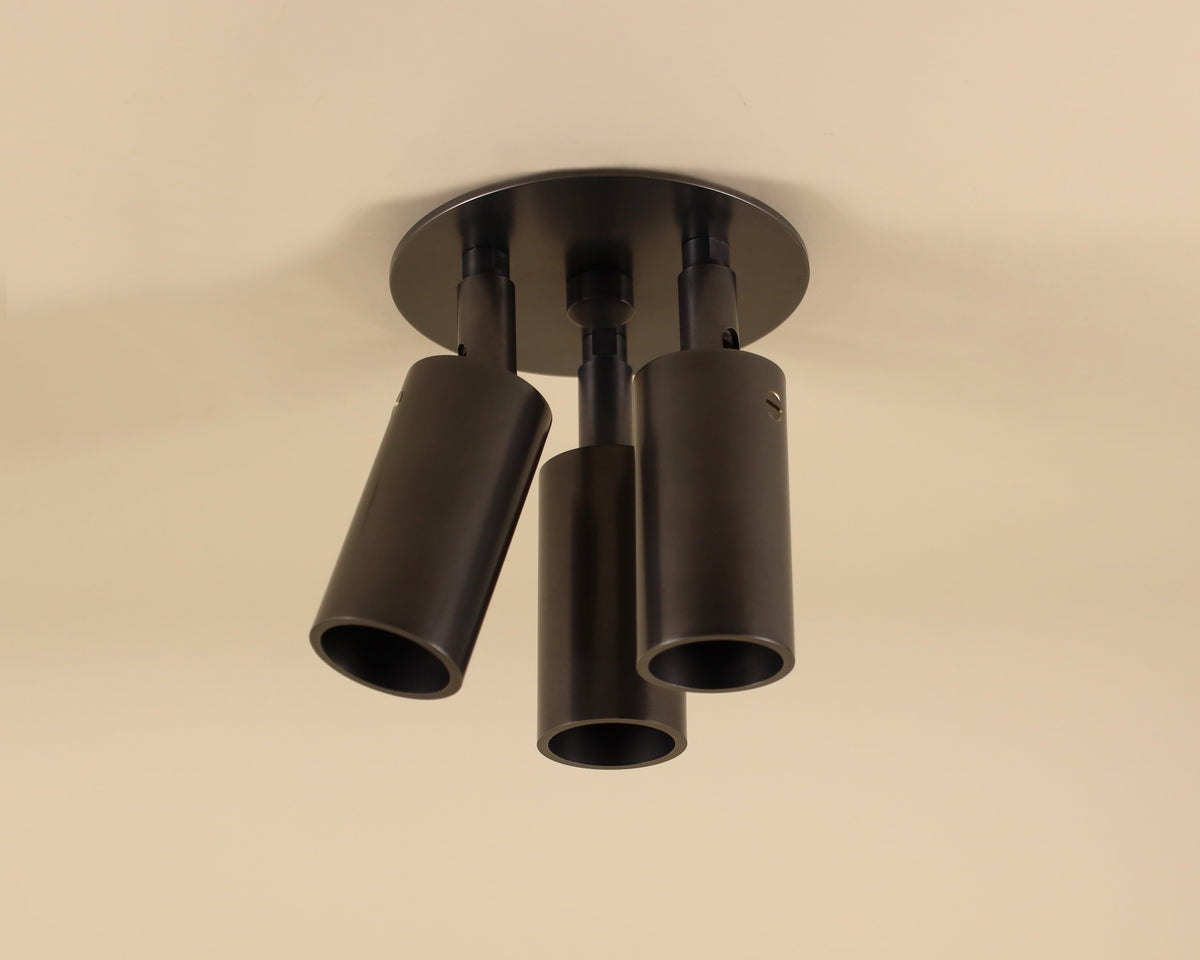 The Long Made Co. Micro Adjustable Spot - Triple is a modern black ultra flush mount fixture featuring three cylindrical, directional LED spot lamps on a round base, shown here against a beige ceiling.