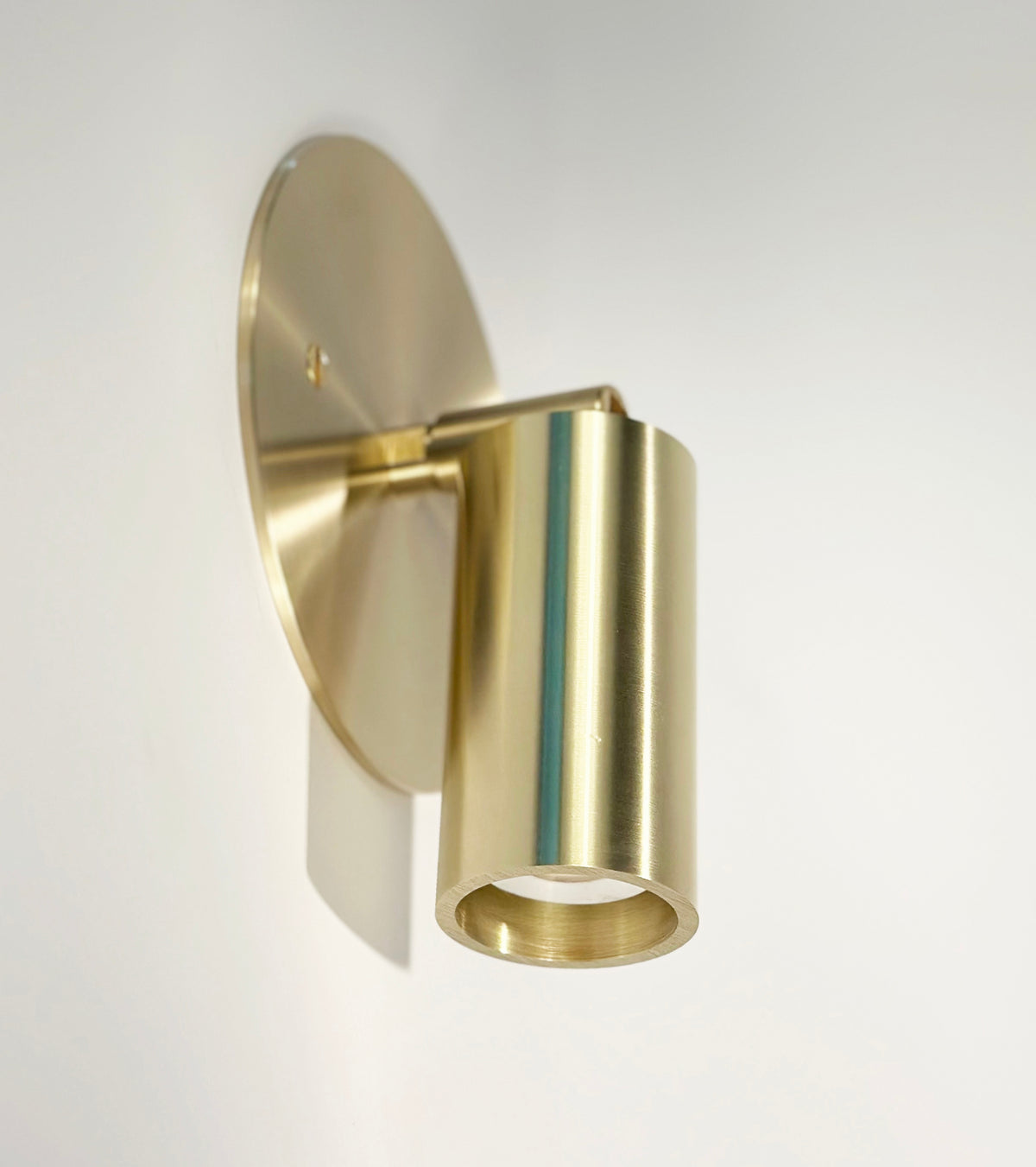 The Micro Wall Spot by Long Made Co. is a modern wall sconce with a cylindrical G9 LED spotlight and round backplate, handcrafted in brushed matte brass for an elegant look on any white wall.