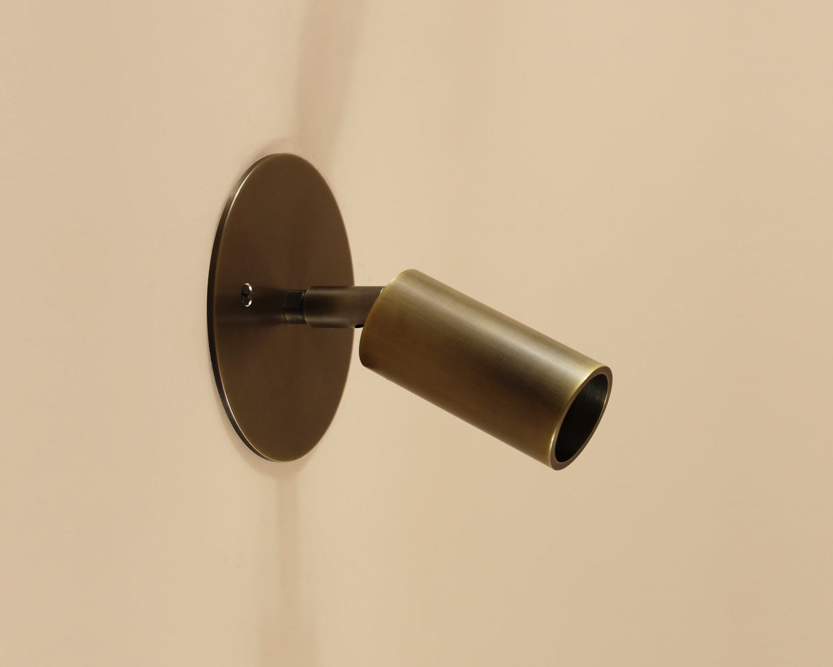 The Micro Wall Spot by Long Made Co. is a modern handmade brass sconce with a cylindrical design on a round base. This ultra-flush fixture mounts against a beige wall and features an LED spot that extends outward, angled slightly downward.