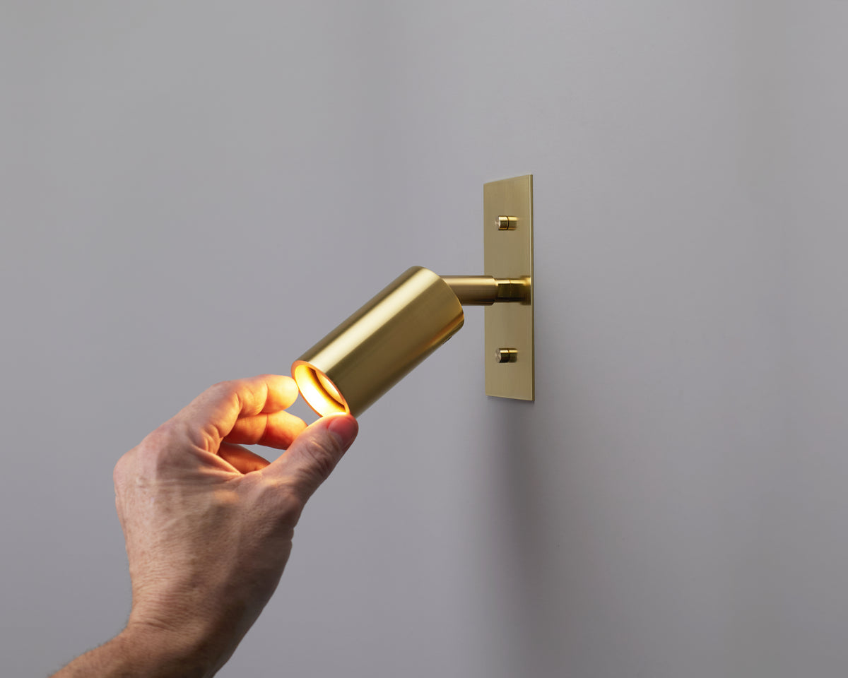 A hand adjusts a Long Made Co. Micro Wall Spot, a modern gold wall-mounted spotlight that is turned on and casts light onto a plain gray wall.