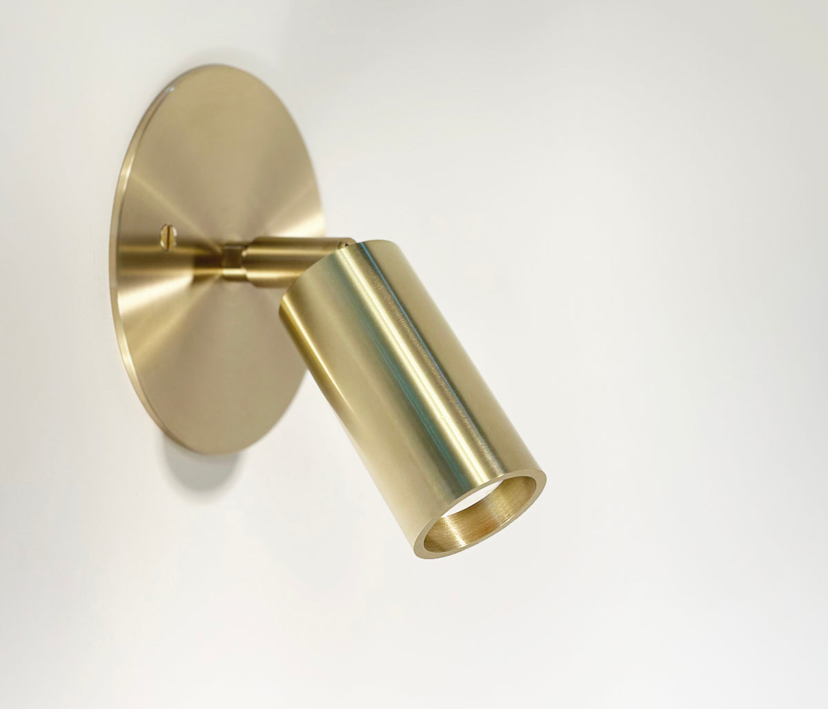The Micro Wall Spot by Long Made Co. is a modern, ultra flush brass wall sconce with a cylindrical design and round backplate, angled downward on a white wall for directional lighting.