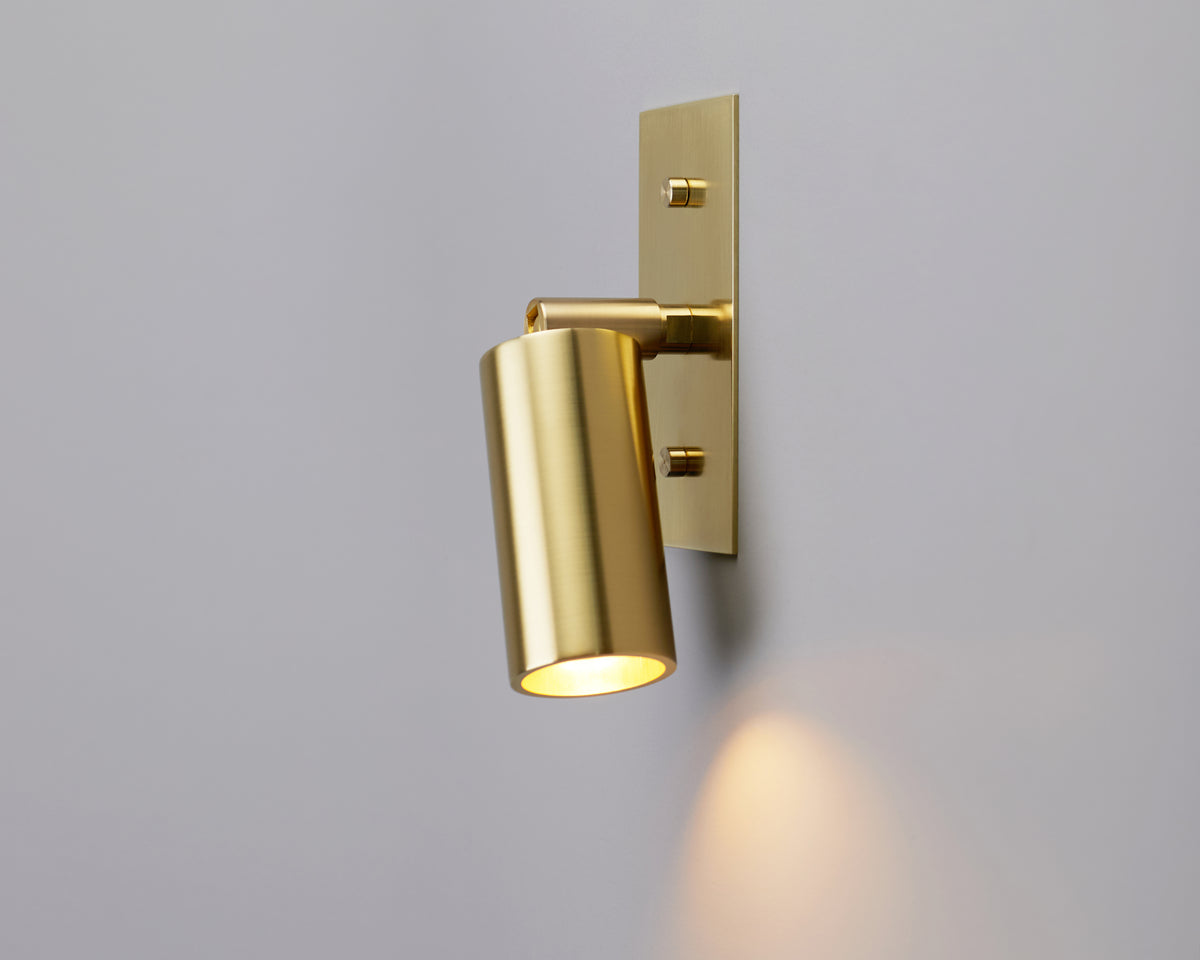 The Micro Wall Spot by Long Made Co., part of the Micro Spot Series, features a modern gold finish with a cylindrical shade and is mounted on a light gray wall, casting a soft, warm downward light—making it an ideal choice for bedside reading.