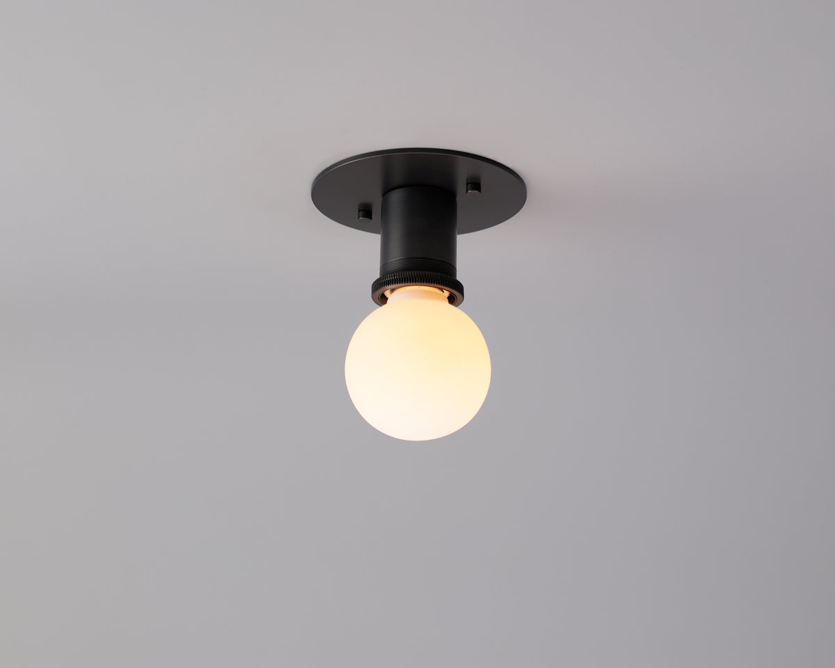 A round, frosted light bulb is mounted on the Brass Minimal Flush fixture by Long Made Co., its simple black design set against a plain gray ceiling and accentuated by the warm elegance of handmade brass.