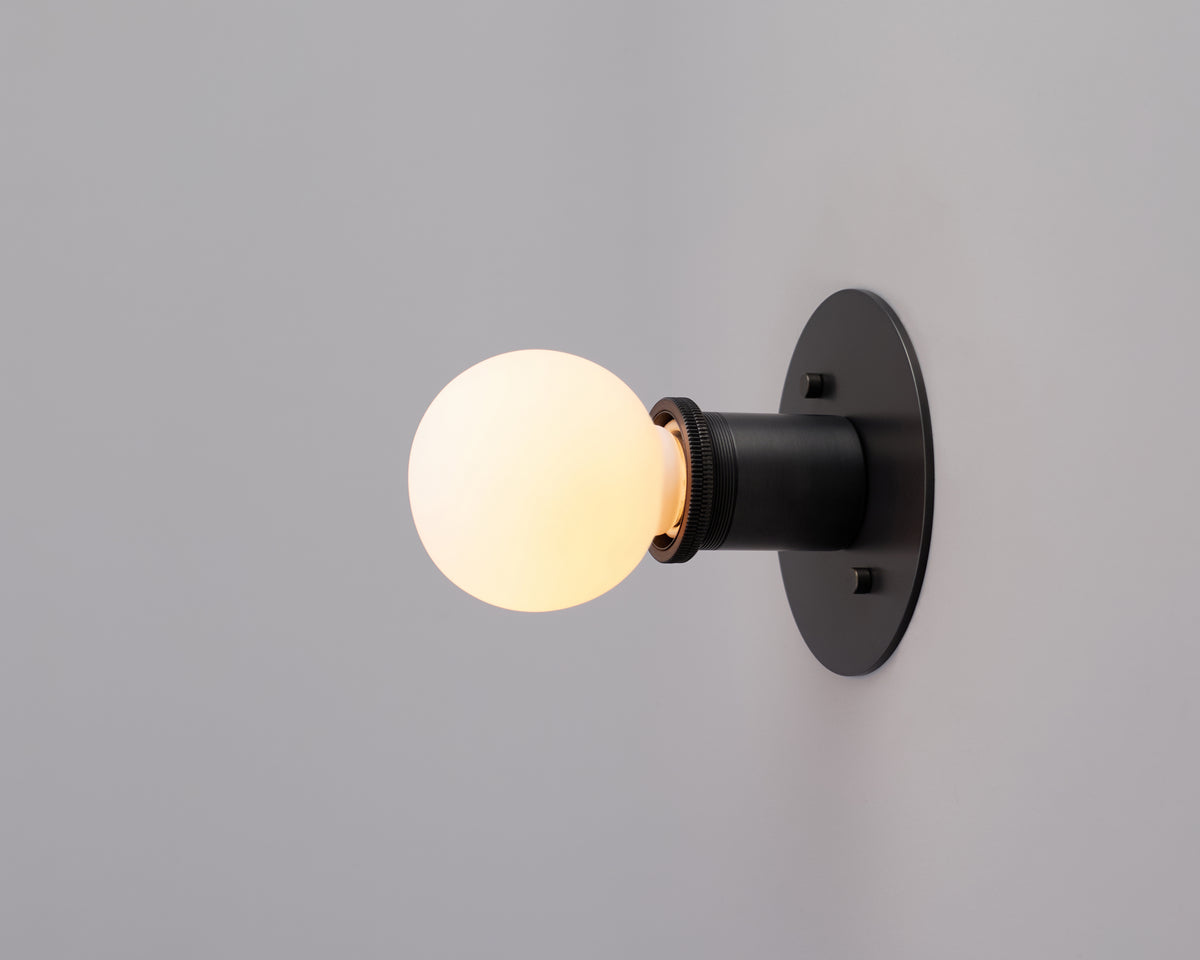 A lit round light bulb is mounted on the Brass Minimal Flush fixture by Long Made Co., set against a plain light gray wall.