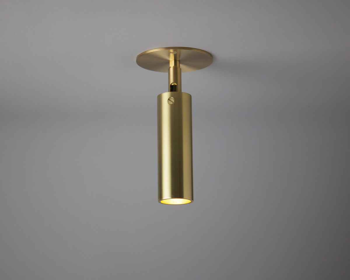 The Micro Adjustable Spot - w/ Long Shade by Long Made Co. is a modern cylindrical brass ceiling light on a flat round base, shining softly downward—ideal for minimalist interiors.