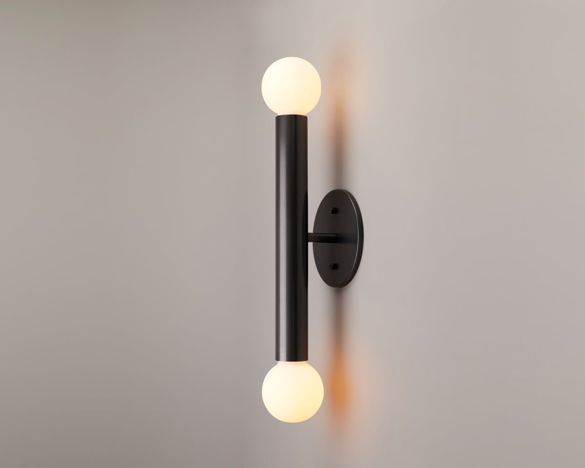 The Miro 2 - 12″ Sconce by Long Made Co. from the Miro Series lighting collection features a modern brass design with a black vertical bar and round white bulbs at both ends, shown mounted on a light gray wall.