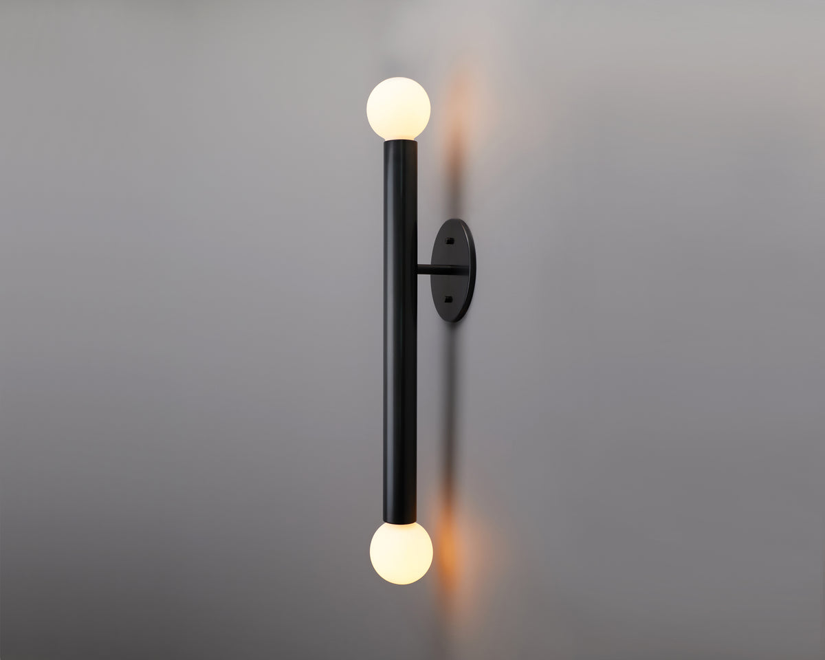 The Long Made Co. Miro 2 XL - 18″ Sconce features a modern brass design with a black vertical rod and round white bulbs on each end, mounted on a gray wall to provide soft upward and downward lighting.