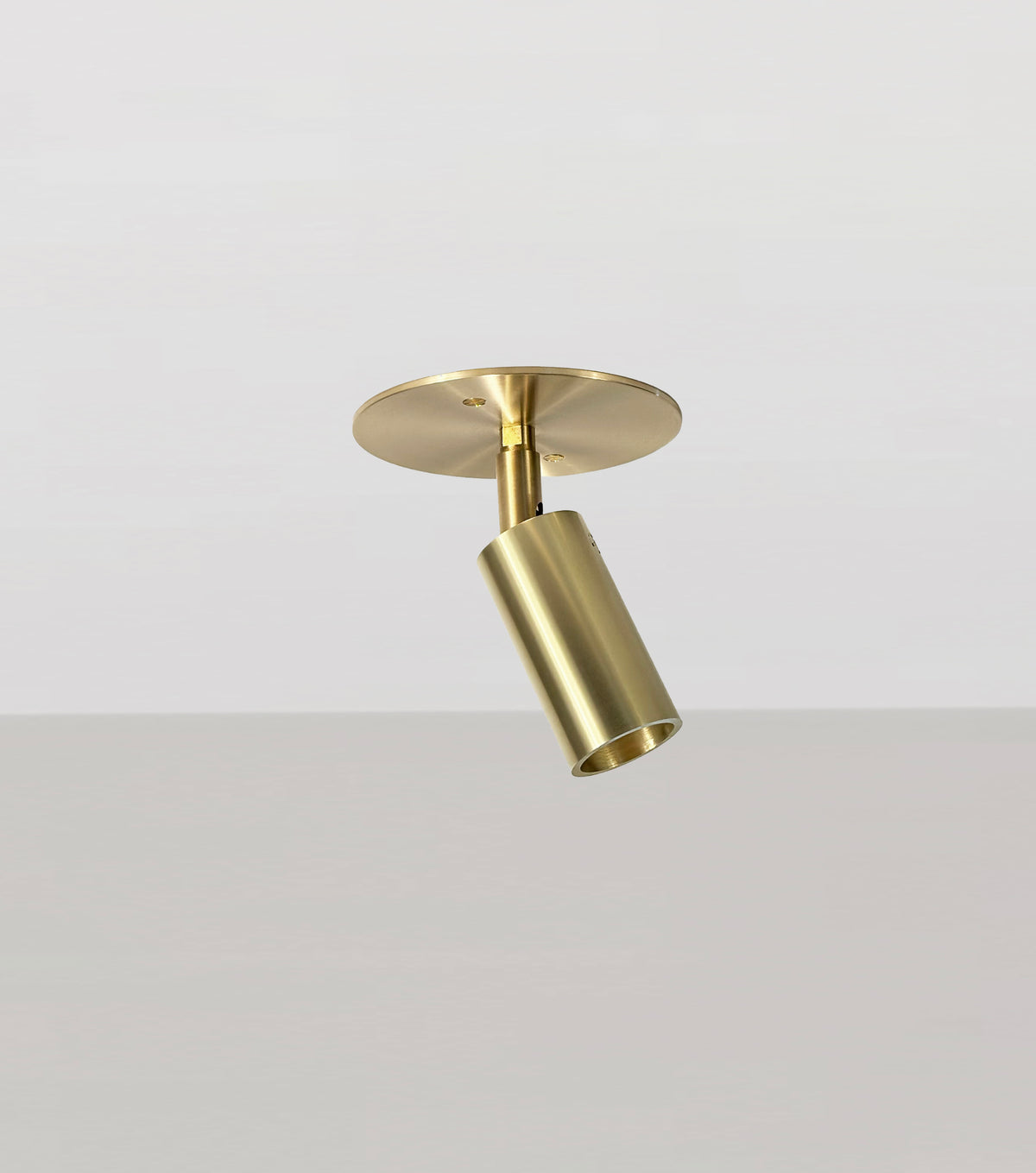 The Long Made Co. Micro Adjustable Spot, a brass-colored cylindrical spotlight with an ultra-thick inset cylinder head, is mounted on a round ceiling plate against a plain light gray background and angled downward.