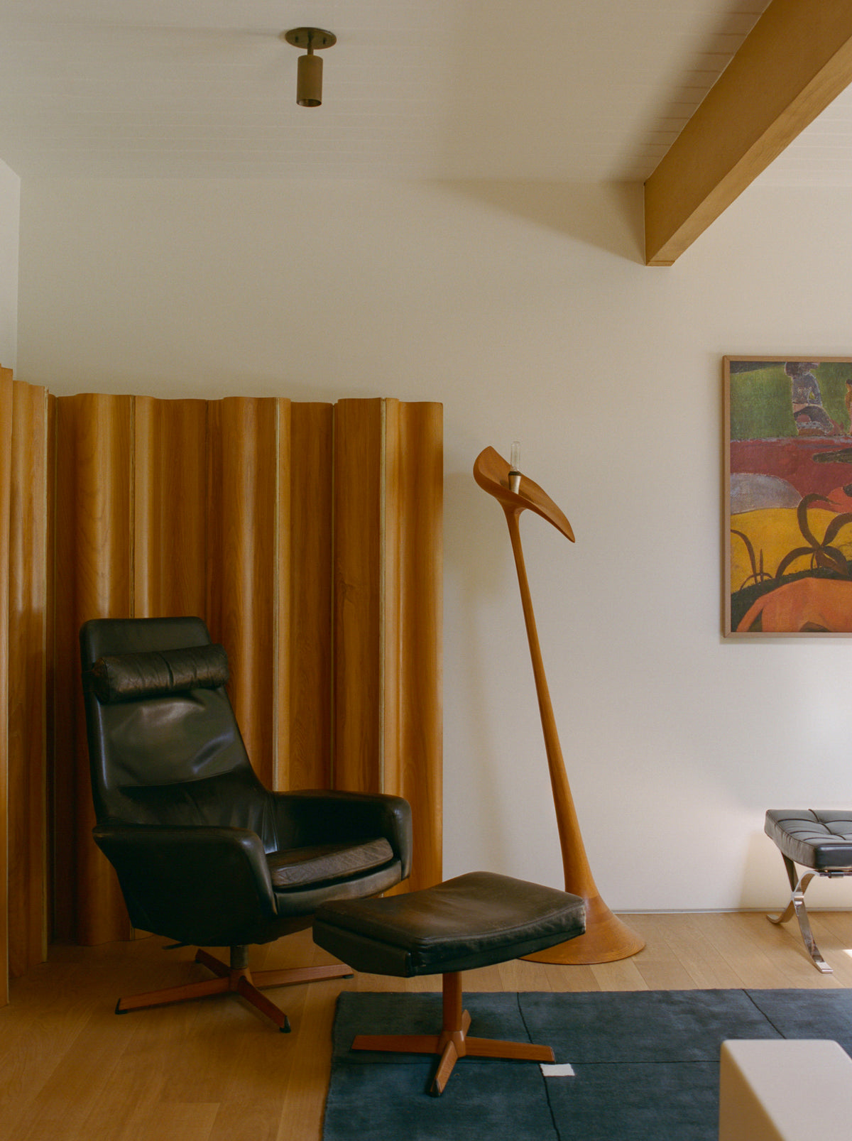 A modern living room corner with a black leather lounge chair, ottoman, wooden divider, sculptural coat rack, colorful abstract art, light wood floors, blue rug, and the Brass Cylinder Adjustable Spot - Top Mount by Long Made Co. for mid-century flair.