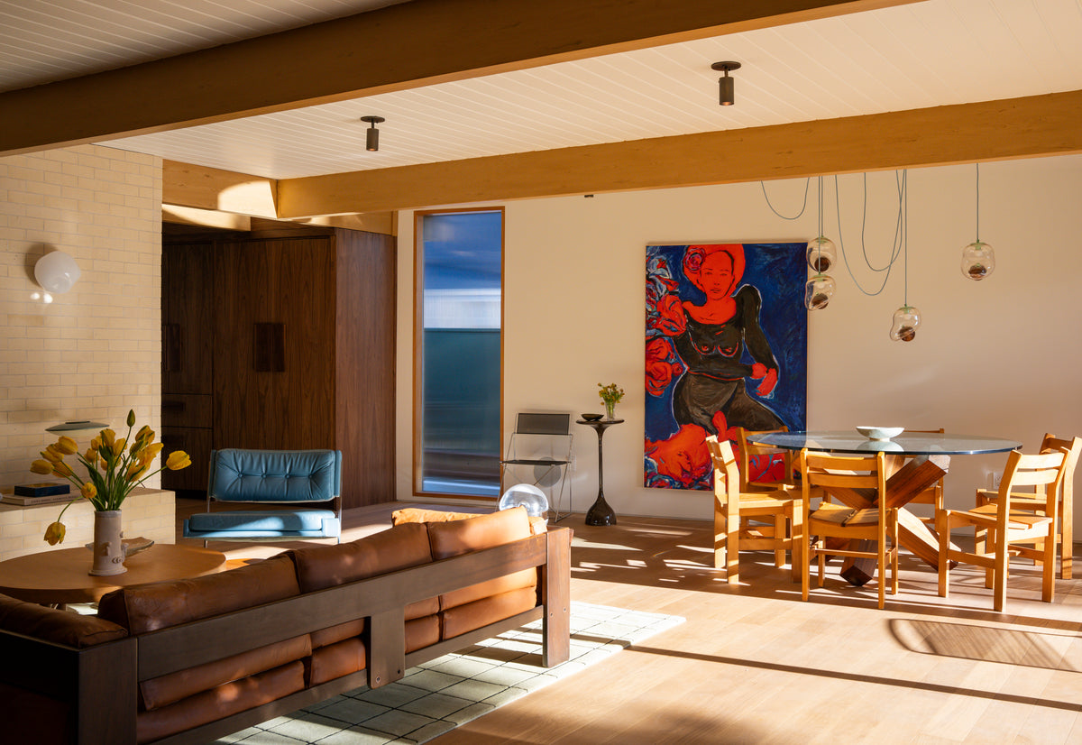A sunlit modern living-dining room with wooden floors, a brown leather sofa, glass dining table with wooden chairs, blue armchair, tulips in a vase, and mid-century lighting featuring the Brass Cylinder Adjustable Spot - Top Mount by Long Made Co.