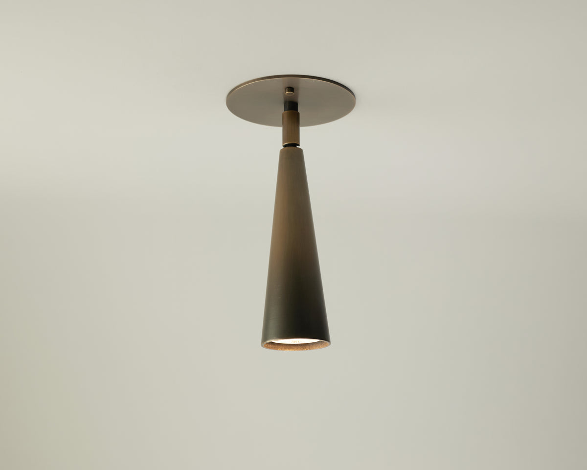 The Pine Adjustable Spot by Long Made Co., a modern black cone-shaped directional ceiling downlight, hangs from a round ceiling mount against a plain white ceiling and wall background, casting a soft downward light.