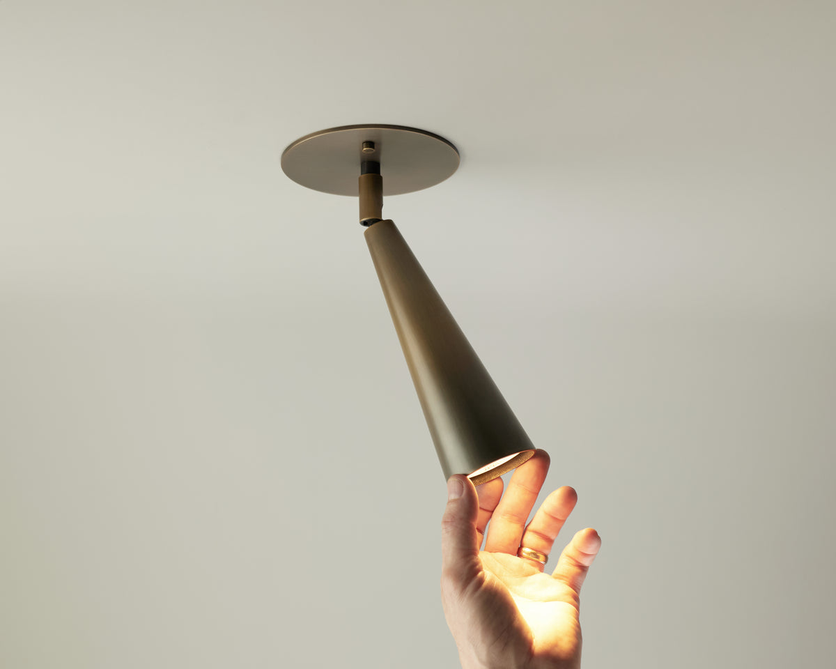 A hand adjusting a Pine Adjustable Spot ceiling light fixture—modern, cone-shaped, and solid brass by Long Made Co., handmade in Texas—with light illuminating the fingers against a plain, light-colored background.