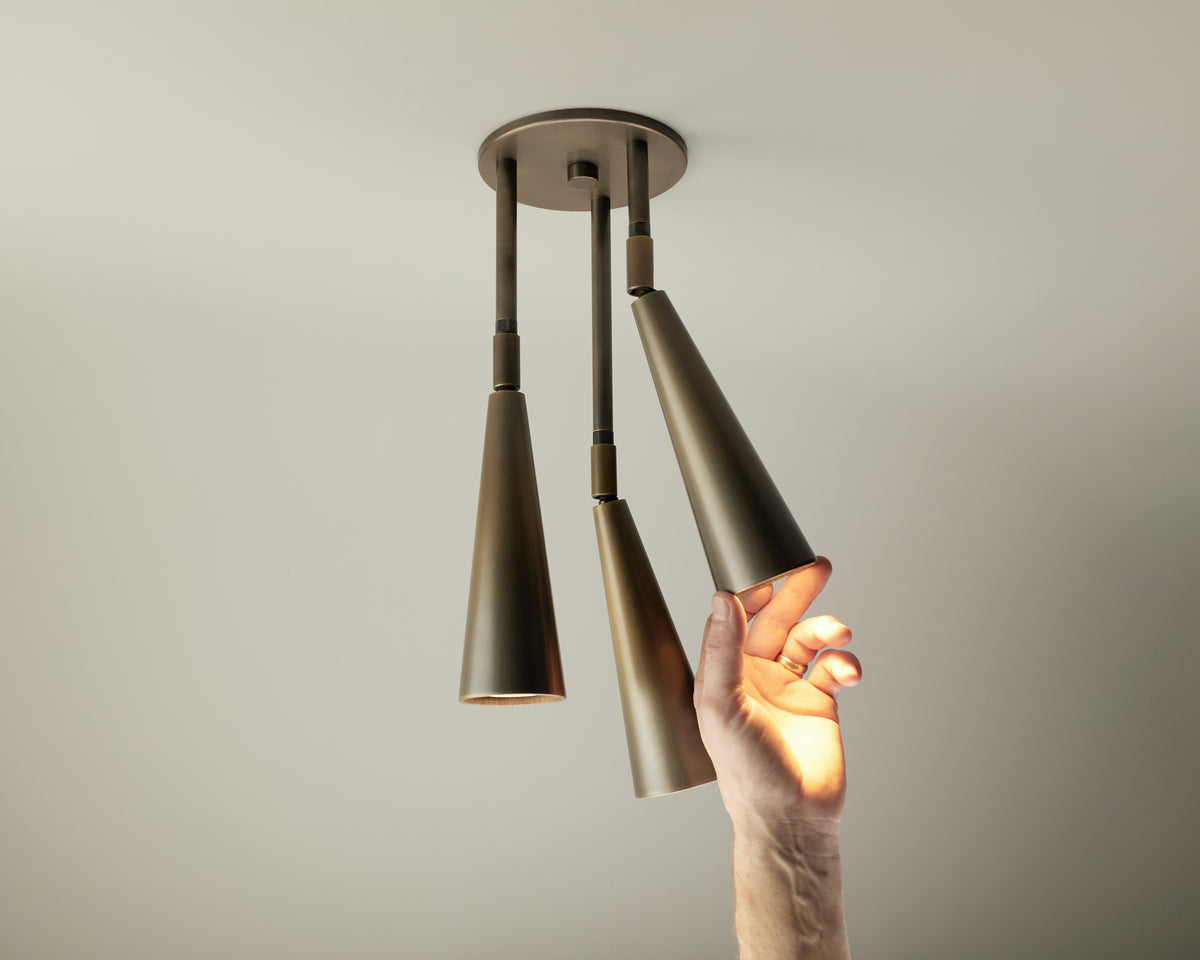 A hand adjusts one of three Pine Cascade Drop Spot lights by Long Made Co.—each an adjustable spotlight—suspended from a solid brass ceiling fixture against a plain, light-colored background.