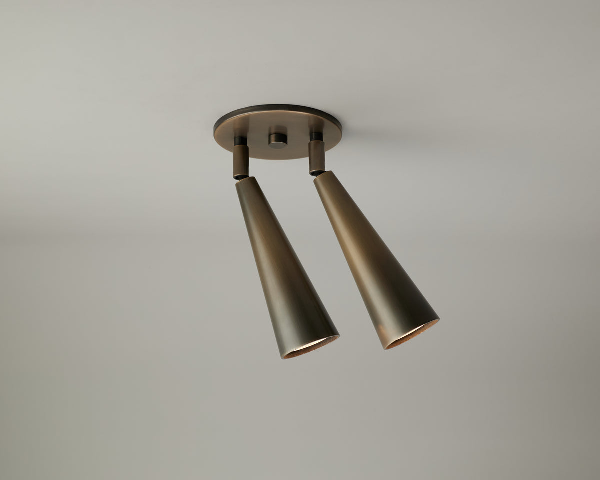 Two bronze Pine Double Spot ceiling spotlights from Long Made Co., featuring a modern, minimalist design, are mounted on a round base against a plain light gray ceiling. Each adjustable spotlight provides versatile illumination, highlighting the timeless appeal of solid brass downlighting.