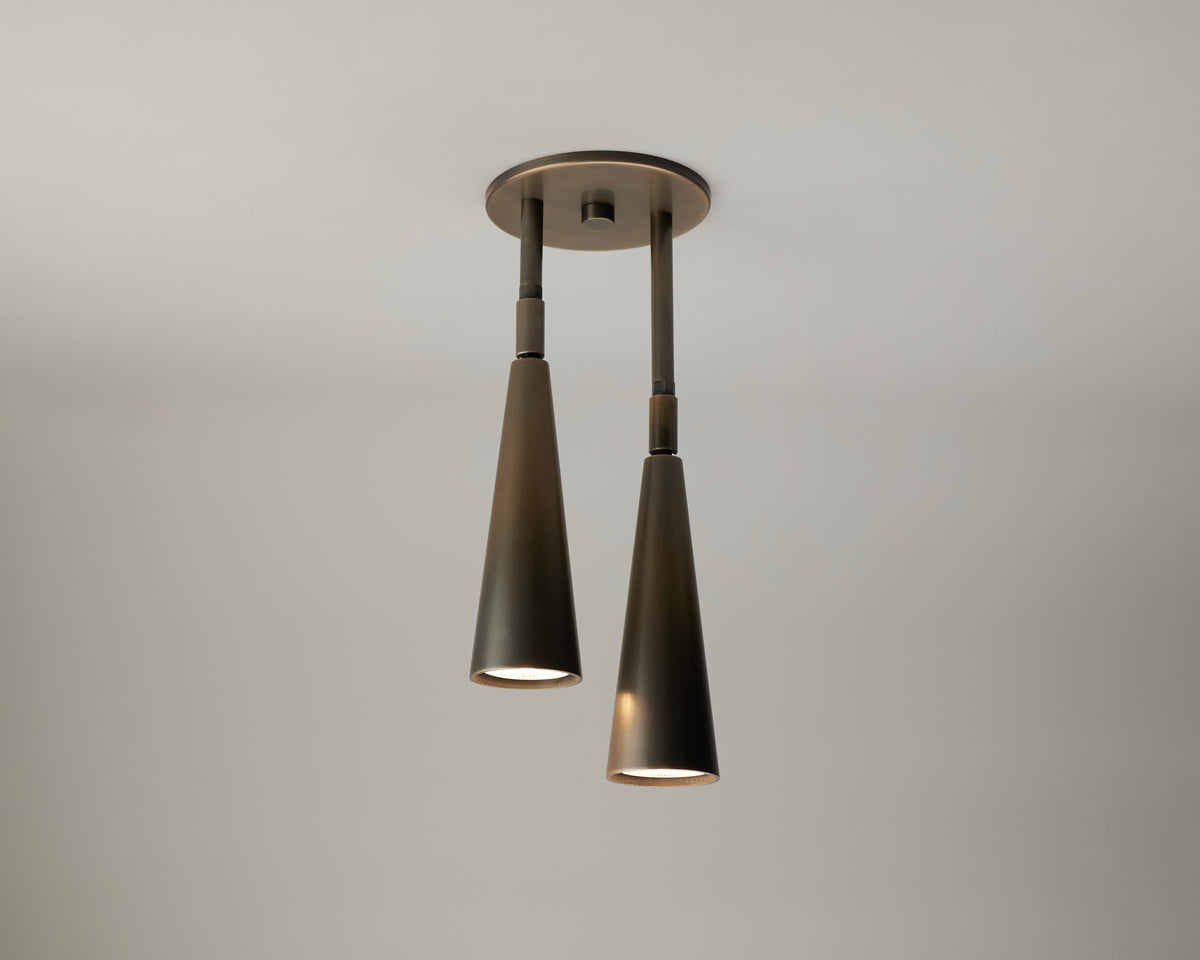 The Pine Double Drop Spot by Long Made Co. is a modern ceiling light fixture featuring two downward-facing, cone-shaped solid brass shades suspended from a circular base, offering elegant versatility—perfect for vaulted ceiling lighting.