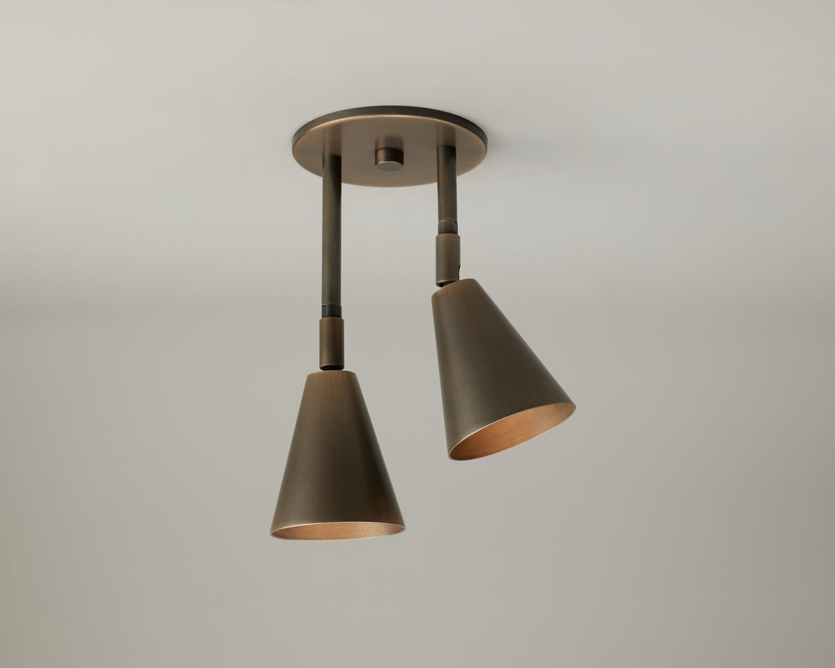 The Tiel Double Drop Spot by Long Made Co. is a modern directional ceiling fixture featuring two downward-facing, cone-shaped metal lampshades attached to a round base and set against a plain light background. Crafted from solid brass lighting elements, this fixture is perfect for vaulted ceilings and brings both functional illumination and elegant style to any contemporary space.