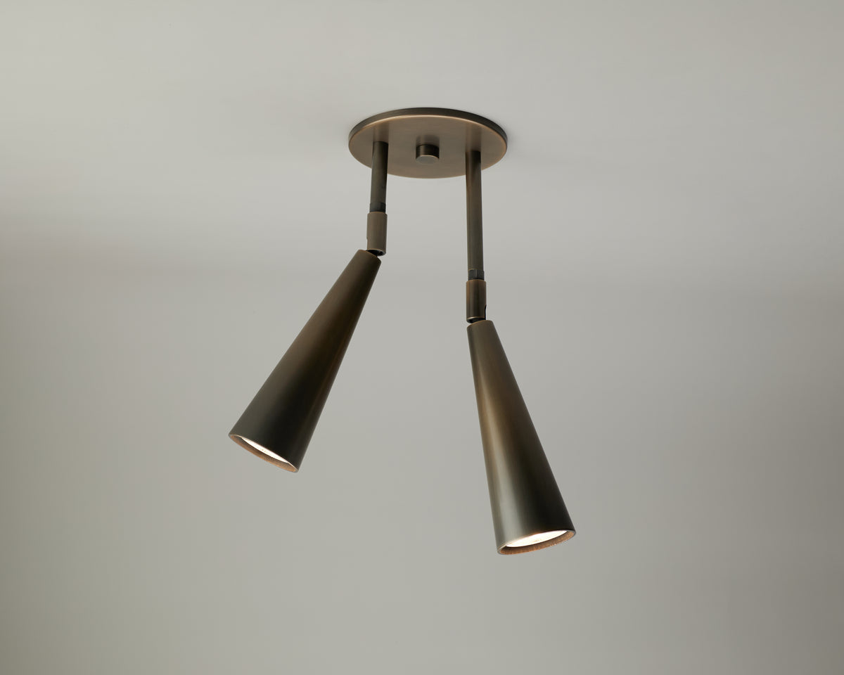 The Pine Double Drop Spot by Long Made Co. is a modern ceiling light fixture with two adjustable spot head downlights, featuring cone-shaped metal shades and mounted on a round base against a plain, light gray ceiling.