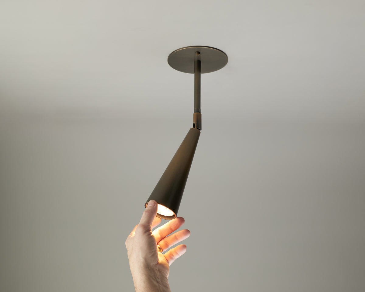 A hand reaches up to adjust or touch the Pine Drop Spot, a modern cone-shaped directional ceiling light fixture by Long Made Co., which is turned on against a plain white ceiling and wall background.