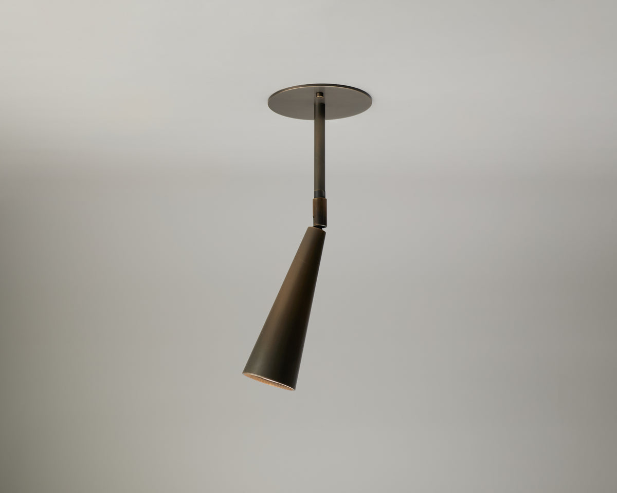 The Pine Drop Spot by Long Made Co. is a modern ceiling light featuring a conical shade, metallic finish, and minimalist design—suspended from a round base against a plain, light-colored ceiling. This directional fixture delivers focused illumination while bringing sleek sophistication to your space.