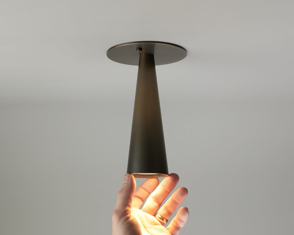 A hand reaches up toward the Pine Flush Spot, a modern cone-shaped brass ceiling light fixture by Long Made Co., with light shining onto the hand.