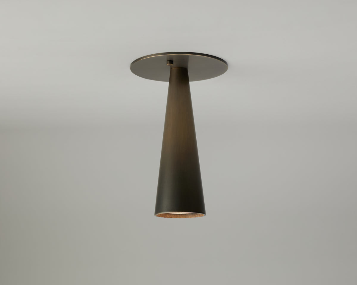 The Pine Flush Spot by Long Made Co. is a modern ceiling light fixture featuring a conical, dark metal shade mounted on a round base against a plain light gray ceiling. This elegant brass ceiling light expertly combines style and reliability as part of Long Made Co.’s handmade lighting collection, and it is fully UL certified for safety.