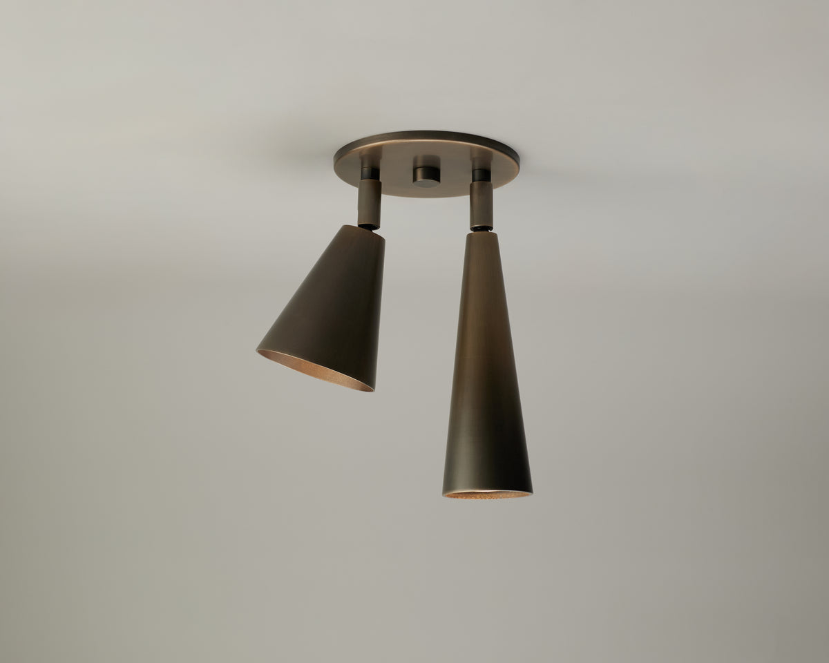 The Tiel / Pine Double Spot by Long Made Co. is a modern ceiling light fixture featuring two cone-shaped, metallic shades angled downward and mounted on a circular base against a plain light-colored ceiling. This adjustable spot light is ideal for adding focused illumination to any space.
