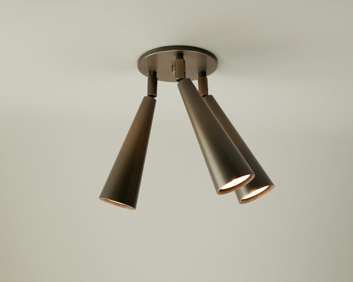 A modern Pine Triple Spot ceiling light fixture by Long Made Co., featuring three adjustable cone-shaped bronze lamps pointing downward, mounted on a circular base against a plain white ceiling.