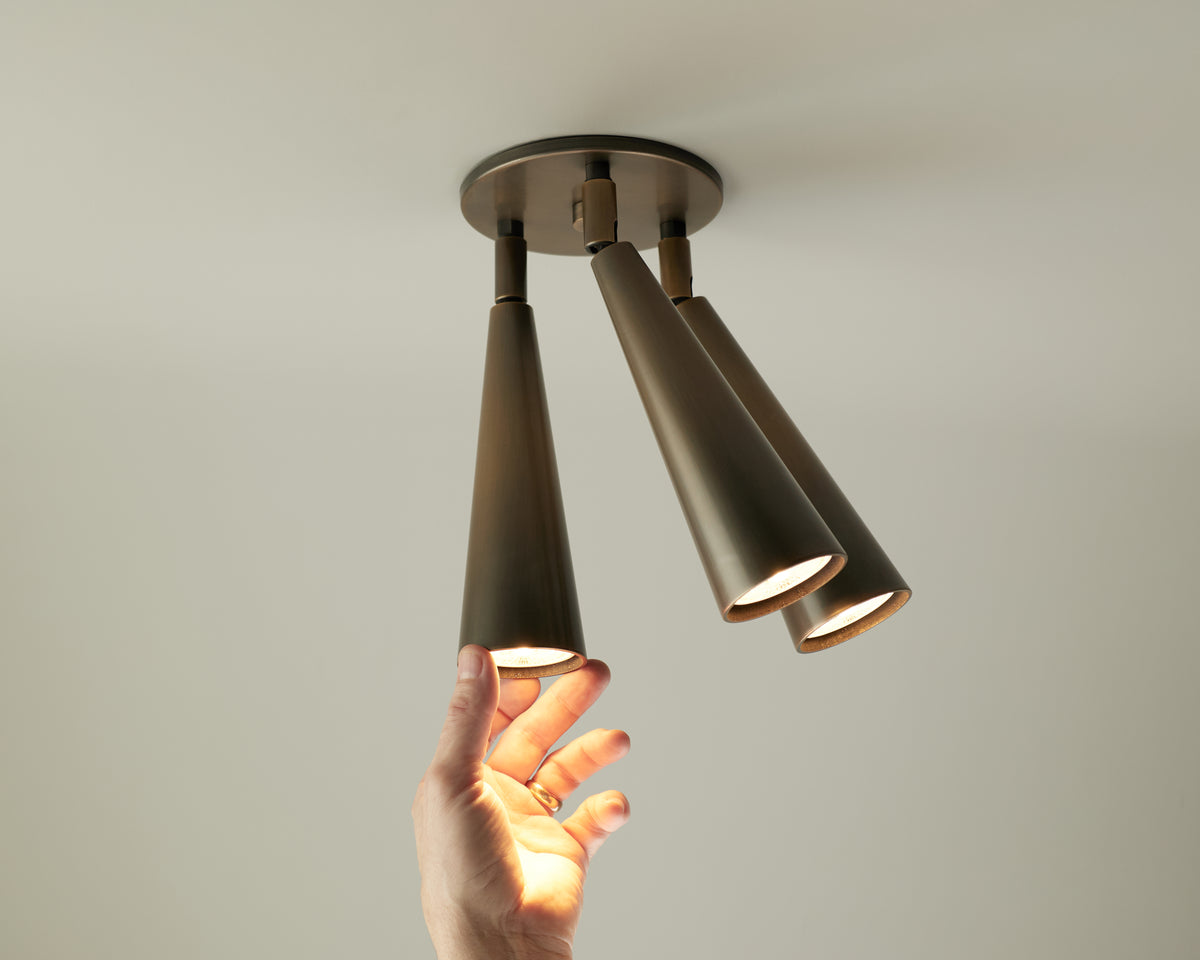A hand adjusts one of three cone-shaped, solid brass ceiling downlights from the Pine Double / Triple Spot by Long Made Co., all illuminated against a plain white ceiling.