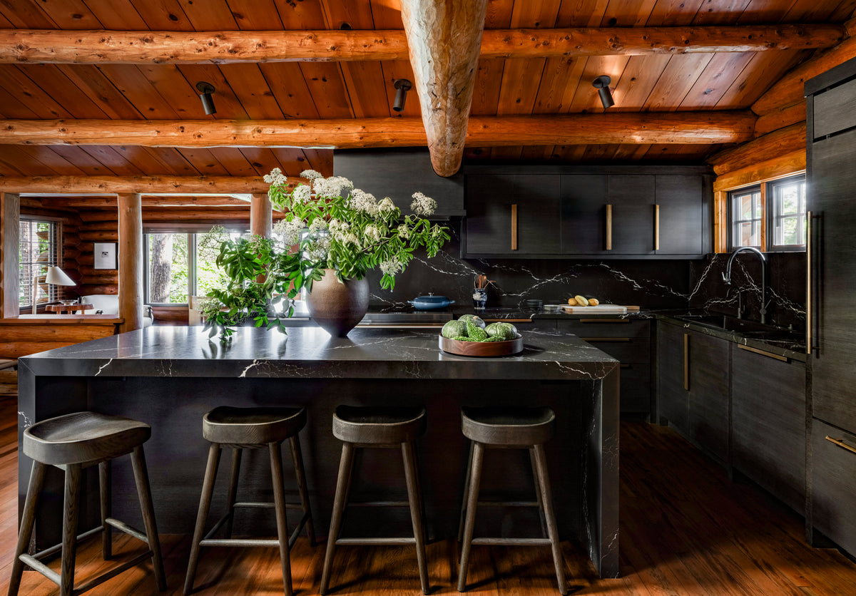 A rustic kitchen with dark wood cabinets and a black marble island is illuminated by Highland Adjustable Spot lights from Long Made Co., highlighting the exposed beams and log walls for a cozy, cabin-like vibe.