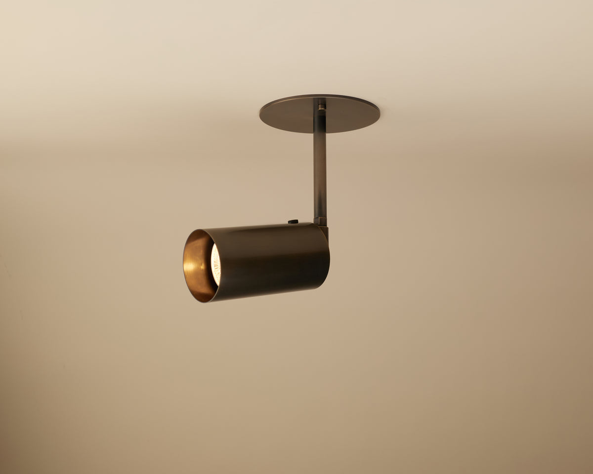 The Long Made Co. Brass Cylinder Drop Spot - Top Mount is a modern ceiling light with a cylindrical brass design, fixed on a round base and angled downward to illuminate the light beige ceiling.