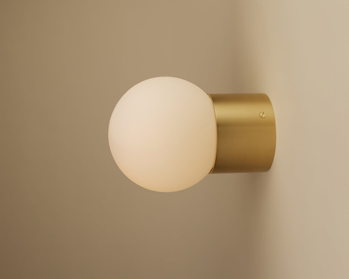 The Beau Laiton Trimless Sphere by Long Made Co. features a frosted glass globe and cylindrical handmade brass base, mounted on a beige wall to emit a warm, soft glow.