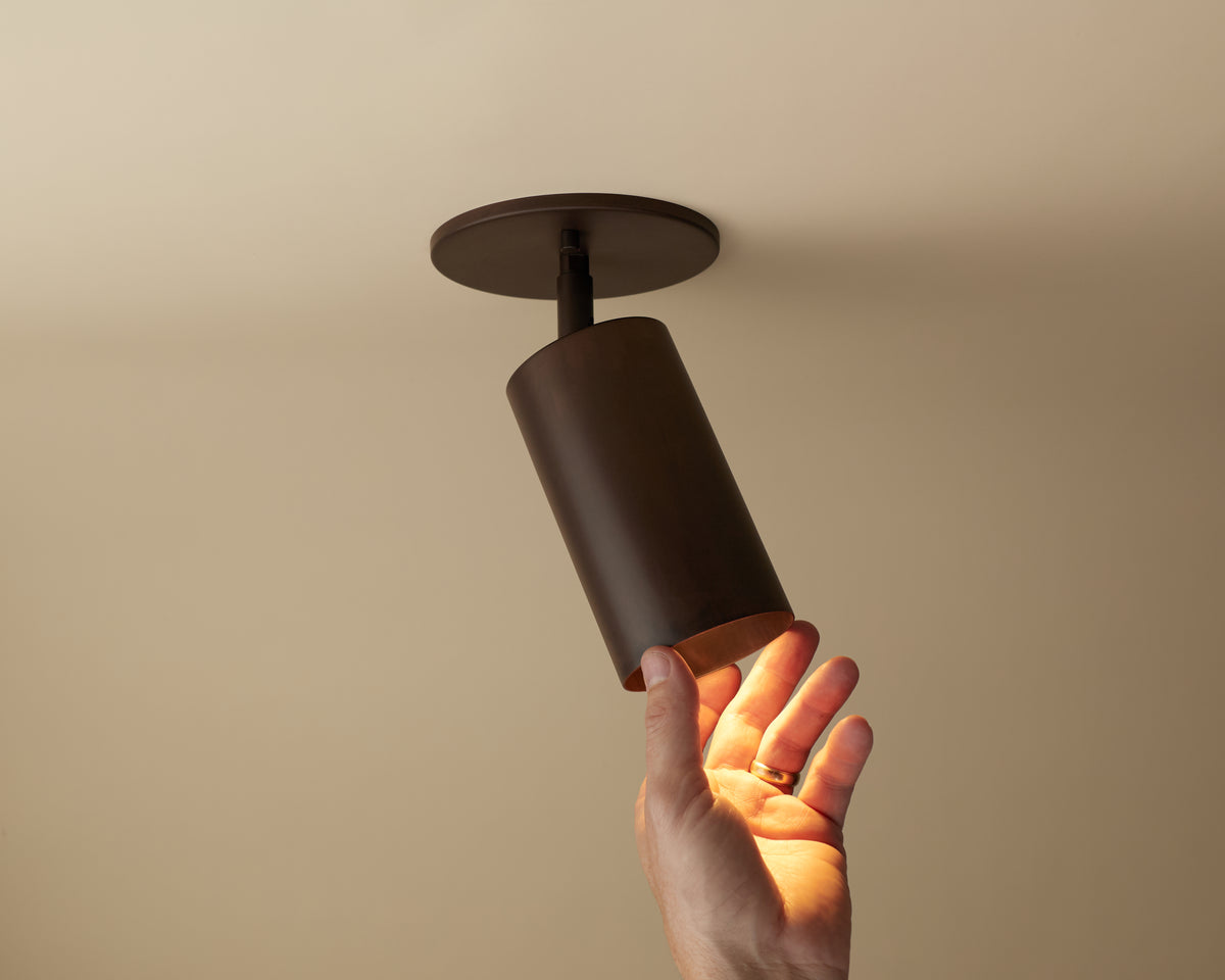 A hand reaches toward the Long Made Co. Cove Adjustable Spot, a modern cylindrical solid steel downlight with a dark metallic finish, mounted on a beige ceiling. The light is on, casting a soft glow.