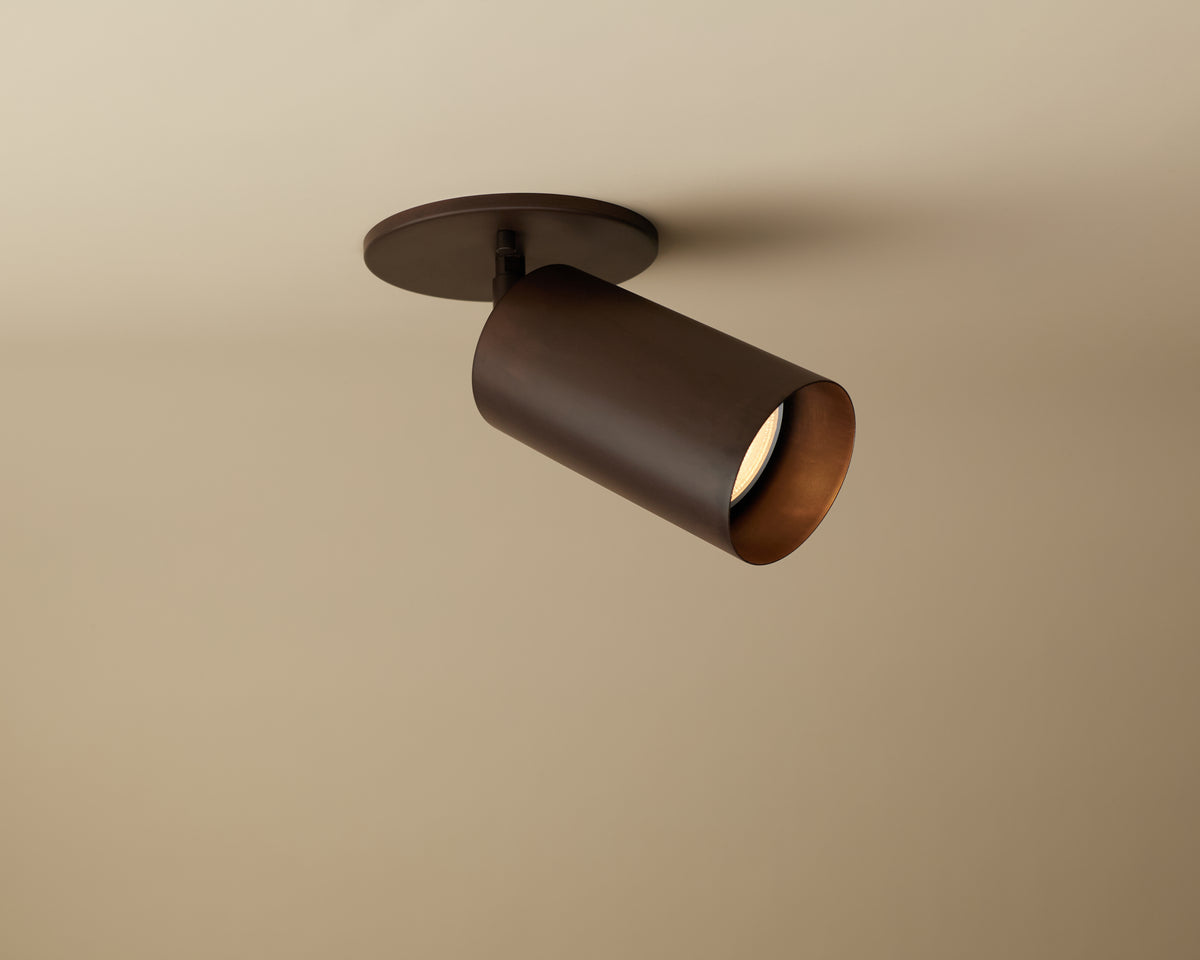 The Long Made Co. Cove Adjustable Spot is a cylindrical brown steel downlight with a round base, mounted on a beige ceiling. It casts a focused downward beam—ideal for minimalist spotlights in custom lighting designs.